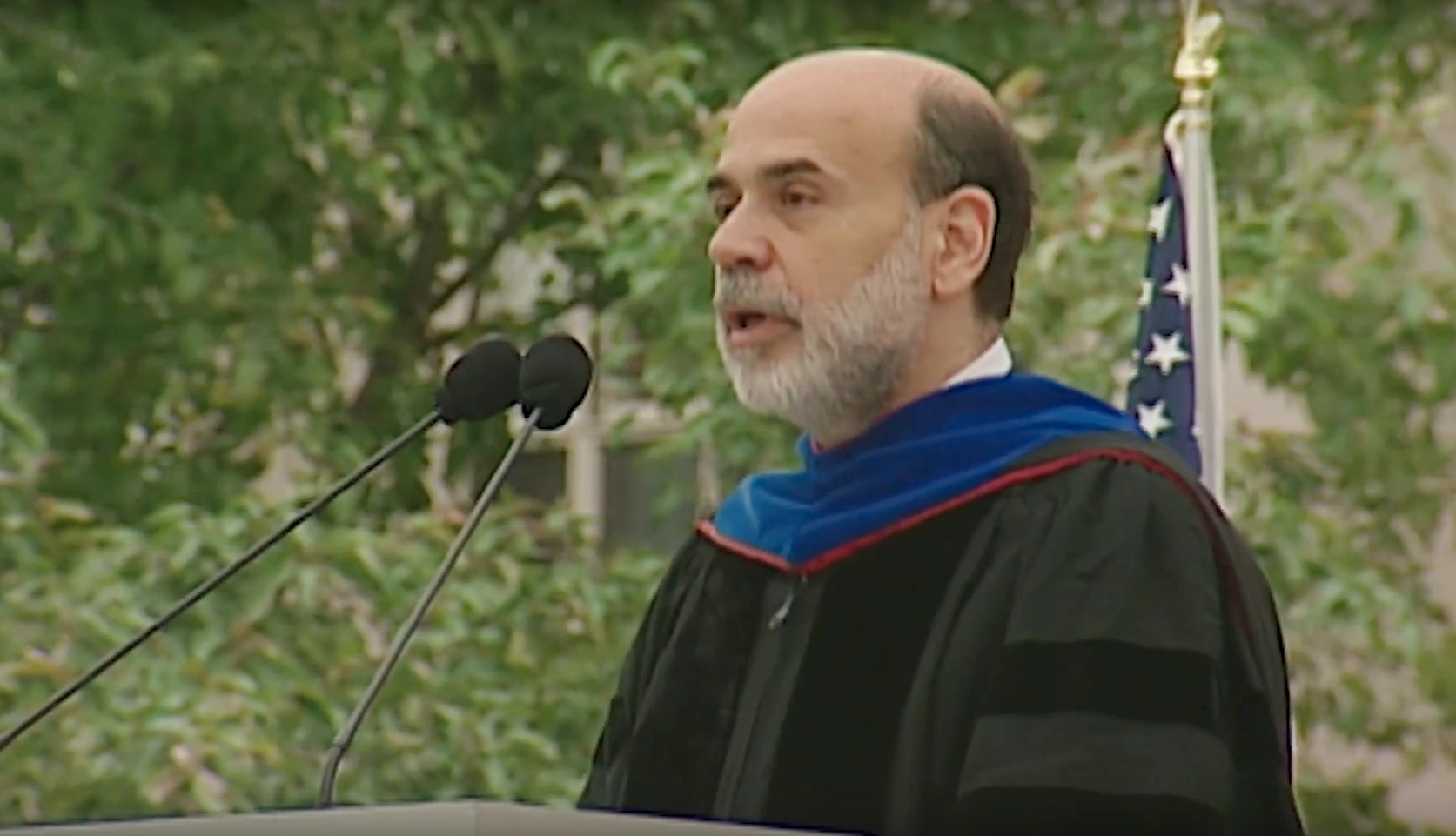 Ben Sholom Bernanke, wearing commencement attire, speaking at podium on outdoor stage, trees in background