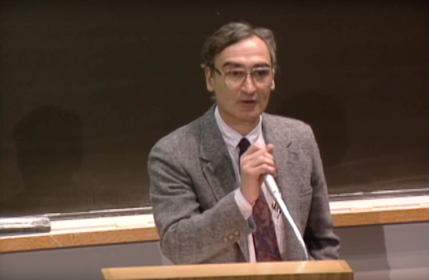 Matthew Meselson speaking into podium microphone in classroom, chalkboard in the background.