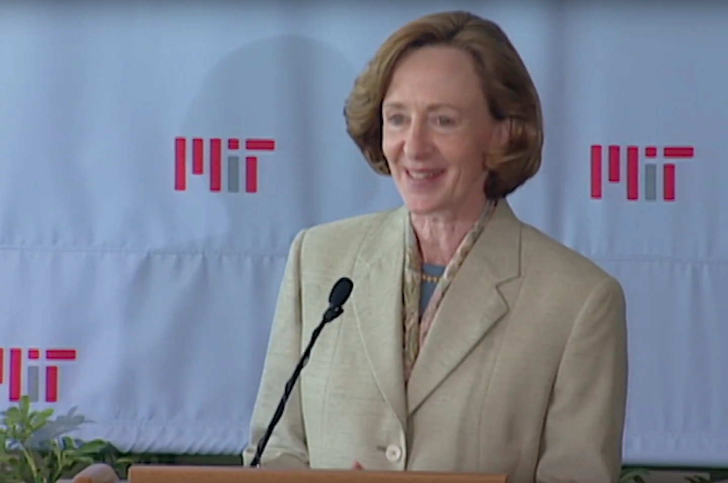 Susan Hockfield speaks at a podium outside, white backdrop with MIT logo in background.