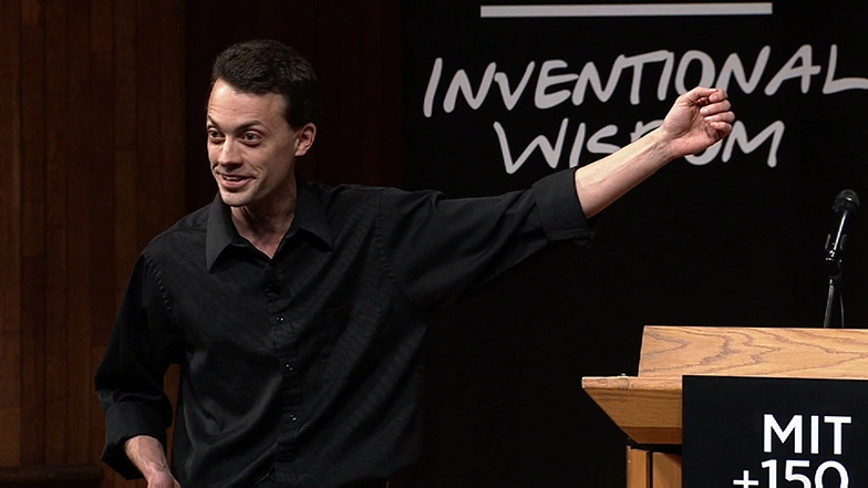 Rob Miller speaking onstage, gesturing with on arm raised, pointing behind him.