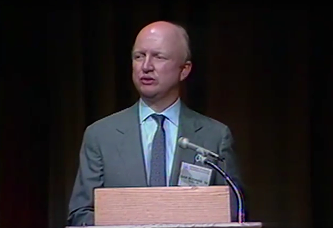 Samuel Bodman stands at podium on stage, black curtain in the background 