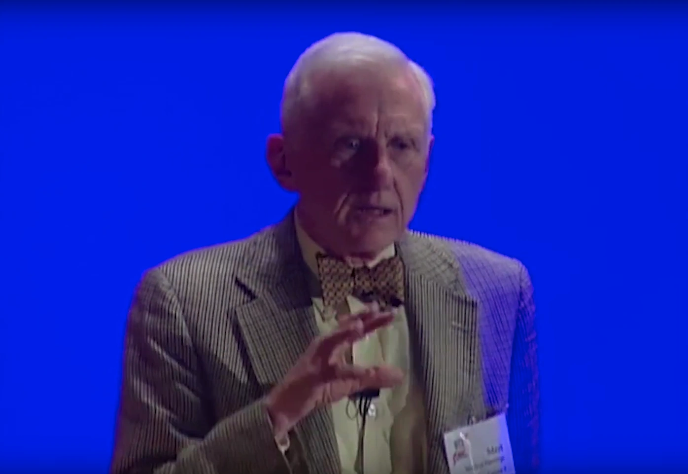 Merton Flemings speaks into podium microphone, dimly lit in front of vibrant blue background