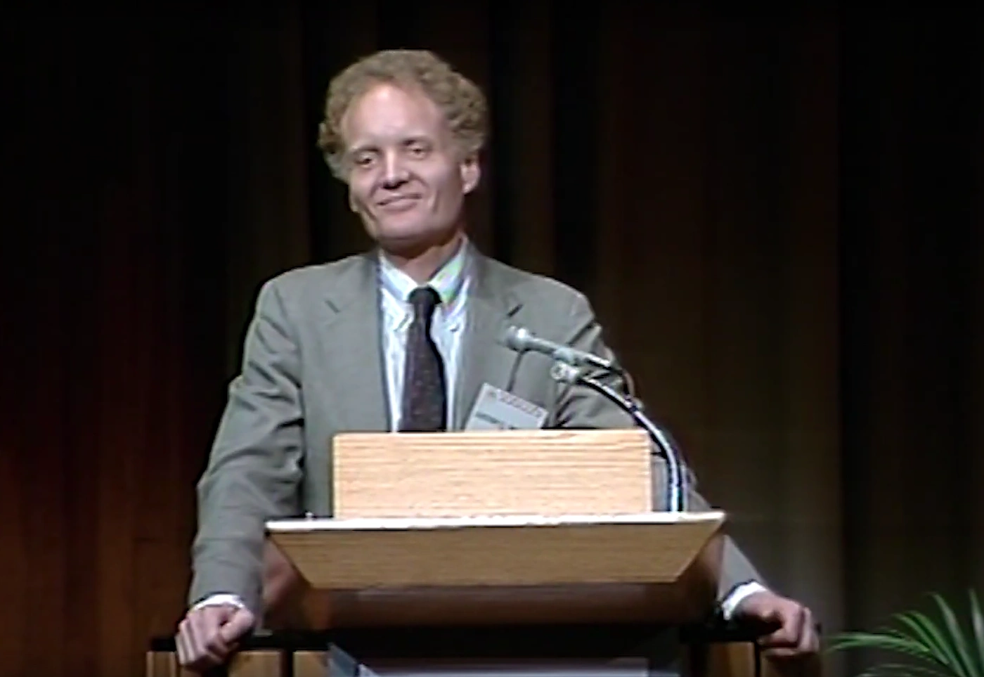 Lester Thurow stands behind podium on stage, black curtain in the background