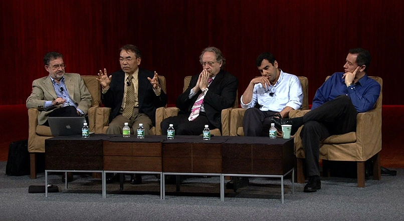 A panel of scholars talk sit in upholstered chairs placed alongside each other on stage, black backdrop