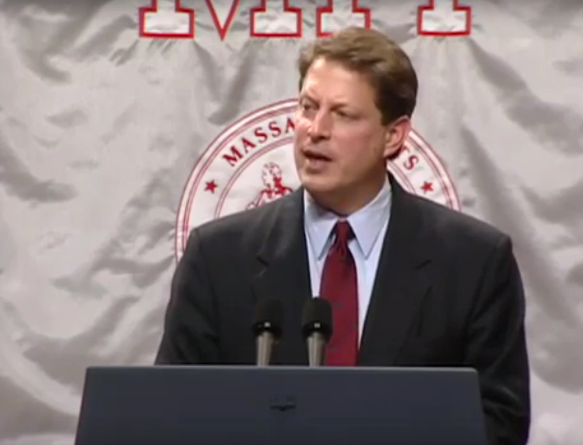 Former Vice President Al Gore giving a speech at a podium, standing in front of a gray MIT flag backdrop