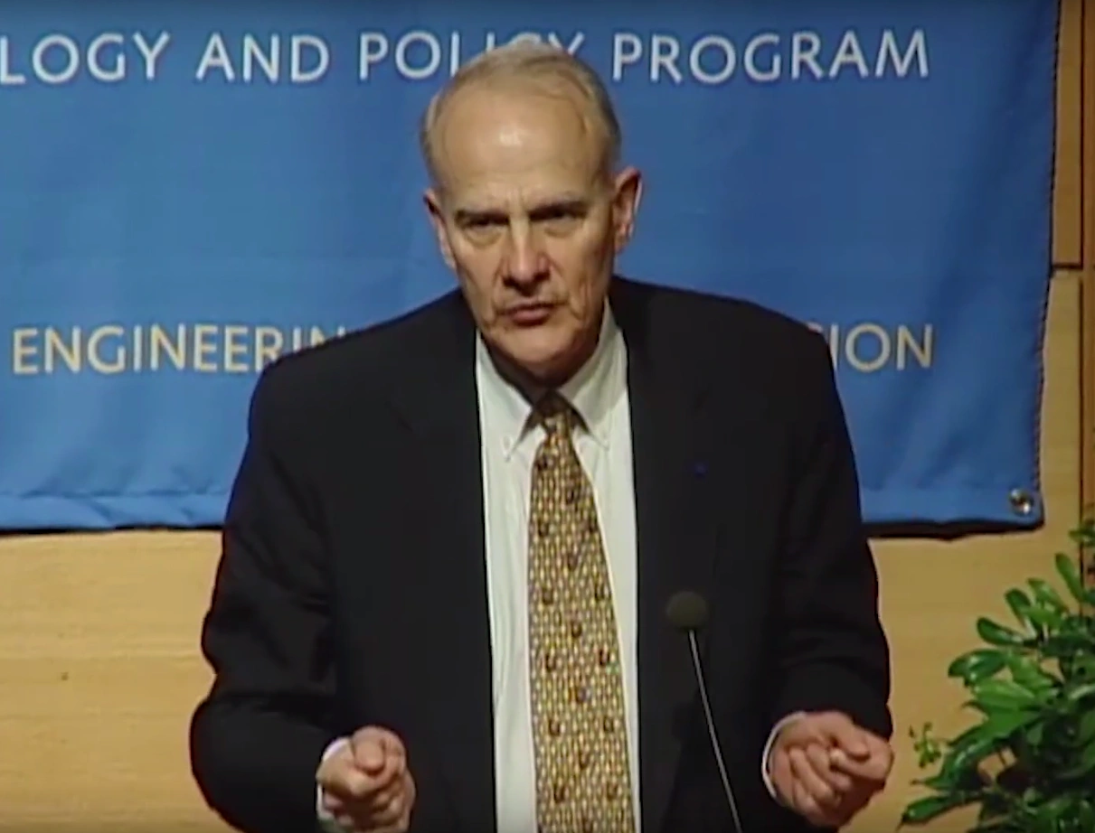 William Wulf speaking at a podium on stage, large blue banner hanging on wood paneled wall with a in the background.