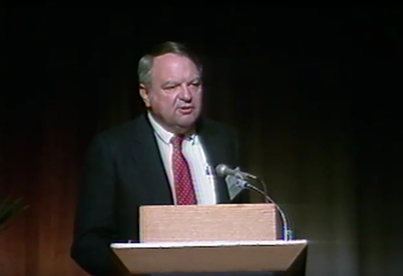 Edward Wilson Merrill stands at a podium on stage, black curtain in the background