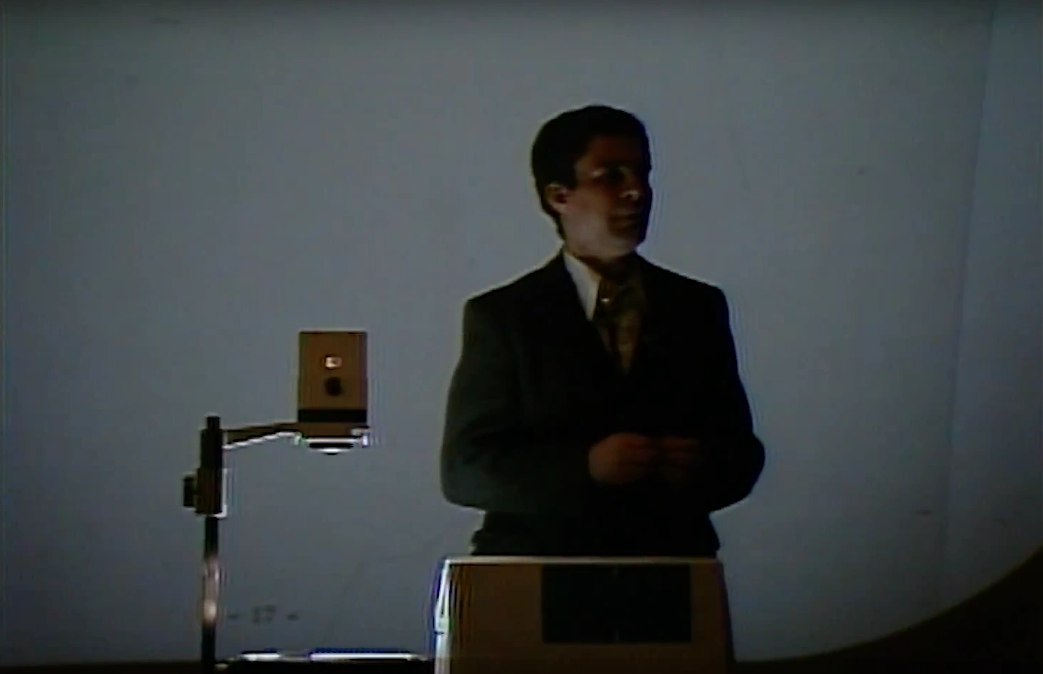 A man stands beside overhead projector with his hands folded in a dimly lit classroom