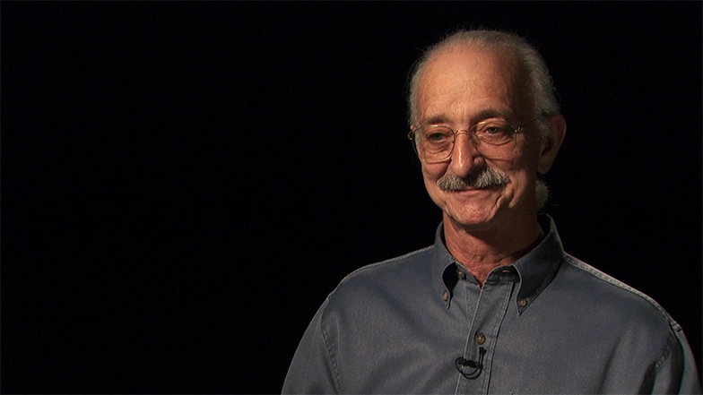 Woodie Flowers seated in front of a black background for an on camera interview, smiling.