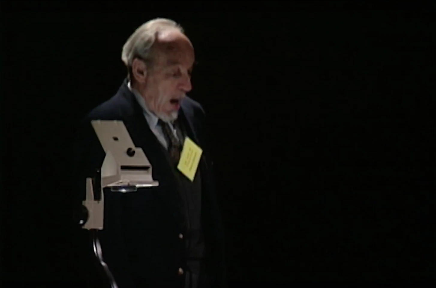Martin Deutsch speaking on stage beside an optical microscope, in front of a dark background.