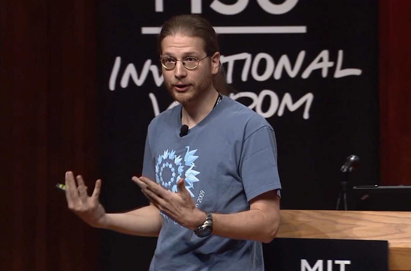 Erik Demaine giving an impassioned speech on stage, a dark backdrop with white text from the MIT150 event in the background.