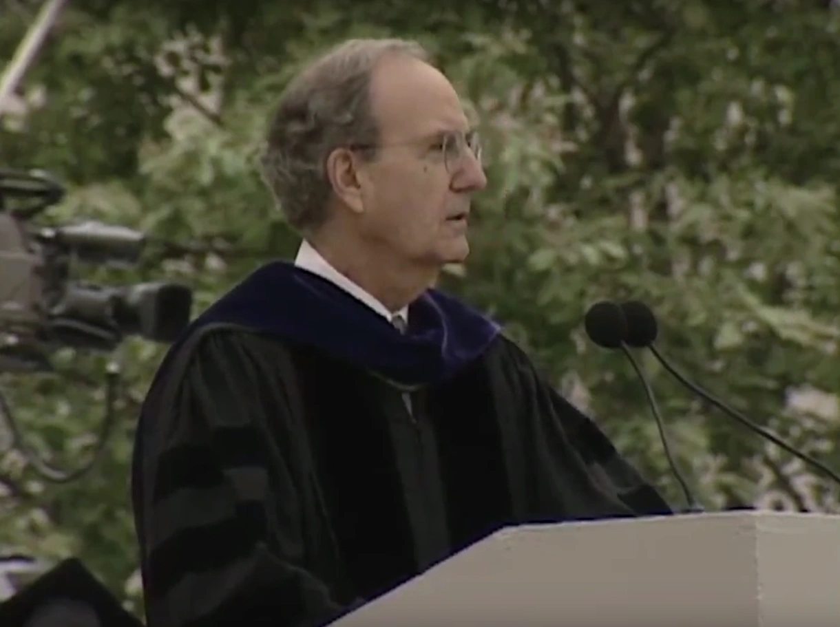 George J. Mitchell Jr., wearing commencement attire, speaking at podium on outdoor stage, trees in background