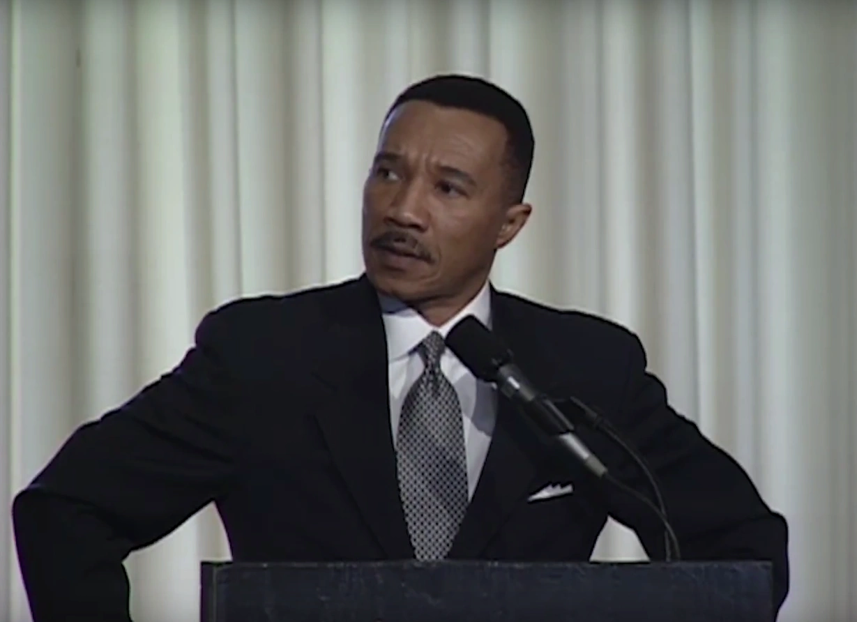 Kweisi Mfume speaking at podium on stage, hands on hips, cream curtain in background