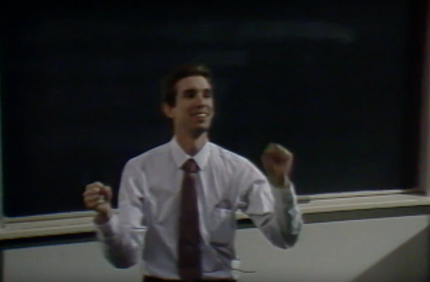 A man expressively lectures in classroom, clenched firsts raised by his shoulders, projector screen and chalkboard in background