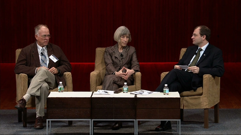 Three panelists have discussion seated in chairs around a coffee table on stage with black background.