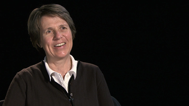 Sallie Chisholm seated in front of a black background for an on camera interview, smiling.
