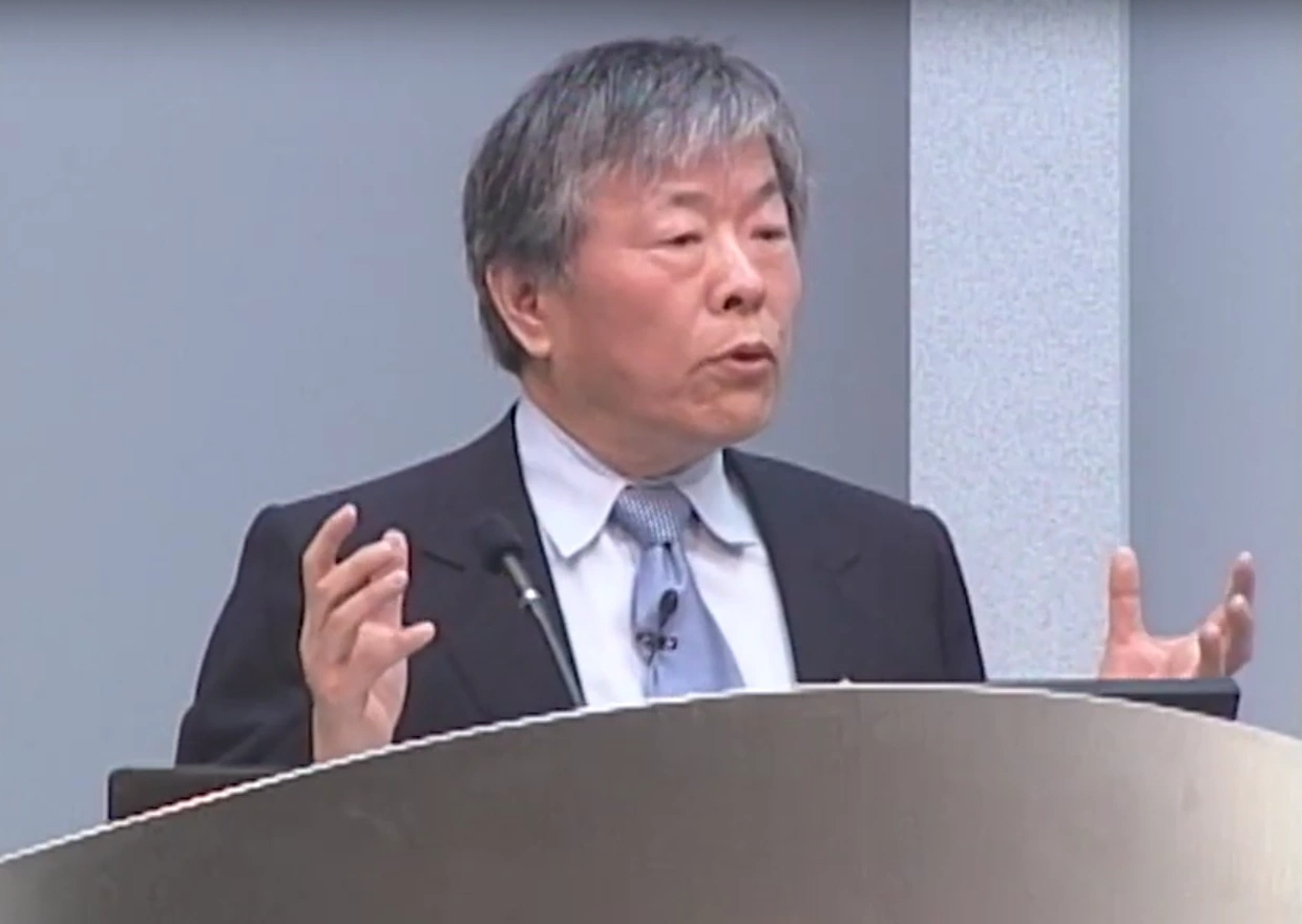 Susumu Tonegawa speaks at a podium in front of a gray background.