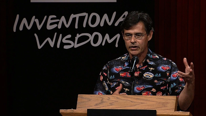 Tony DeRose speaks at a podium onstage, wearing button-up shirt with cartoon cars on it