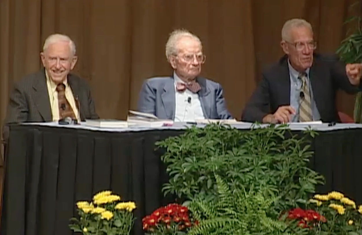 Franco Modigliani, Robert M. Solow, Paul Samuelson, Lawrence S. Bacow sit at panelist table with potted flowers in front.