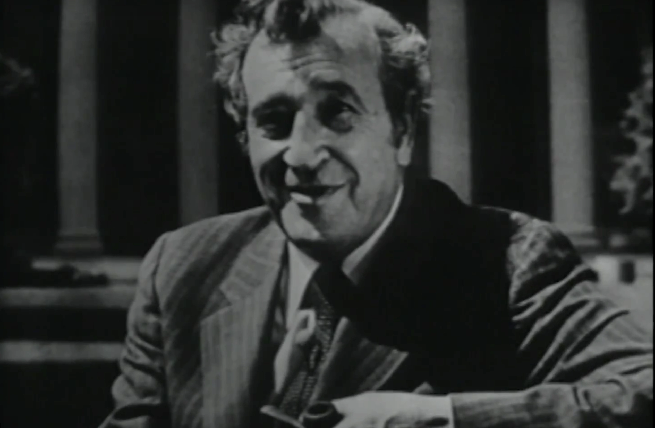Jerome Wiesner standing outdoors, resting his arm on railing and smiling, columns from MIT building in background