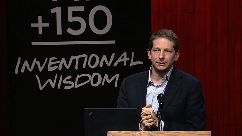 Joshua Tenenbaum speaking at a podium onstage, red curtain in background.