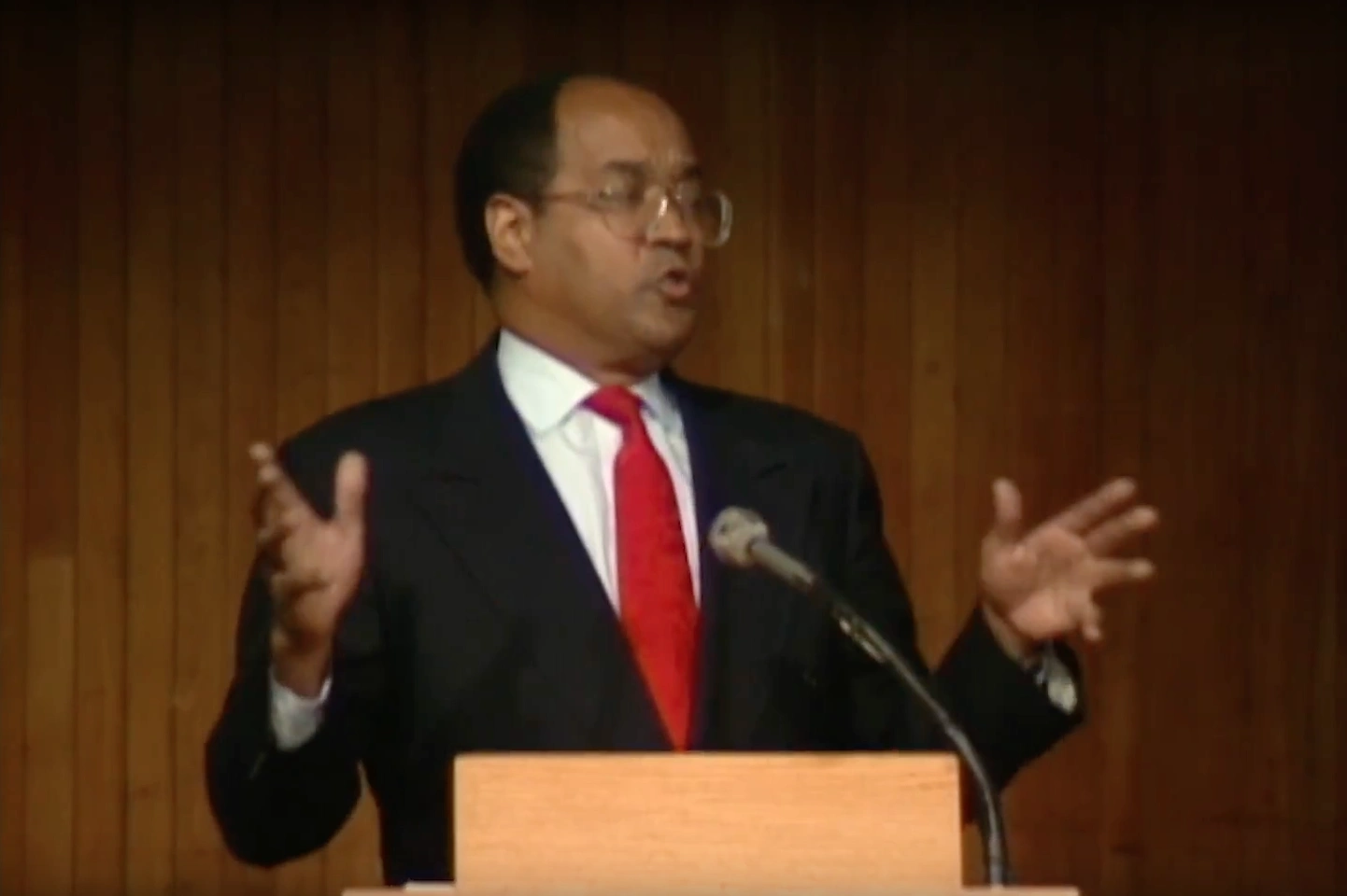 William H. Gray III speaking at a podium on stage, red curtain in background.