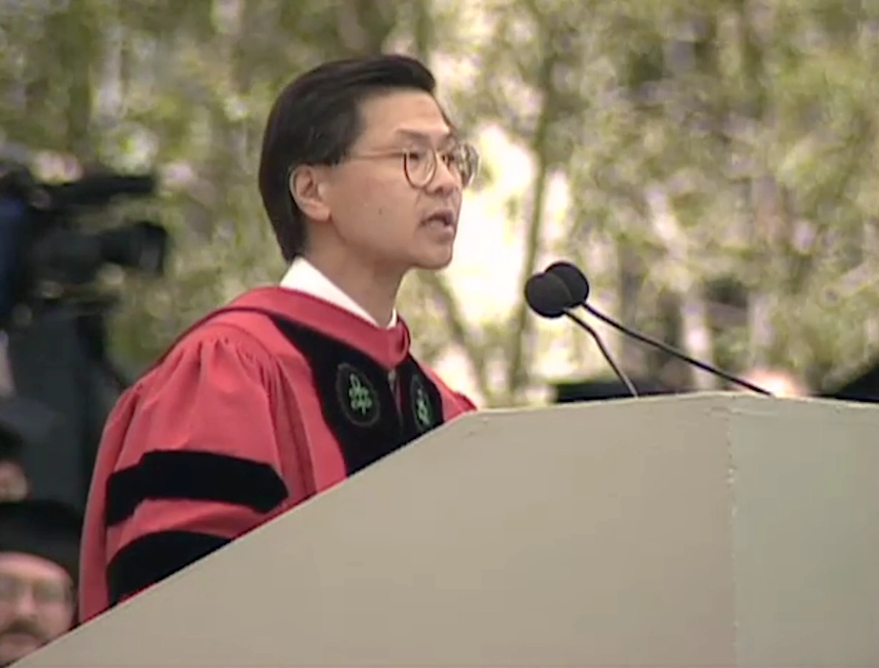 David Ho, wearing commencement attire, speaking at podium on outdoor stage, trees in background