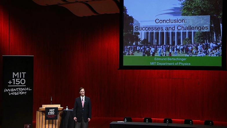 Edmund Berschinger stands beside podium on stage, a giant projector screen and red curtain in backgroud