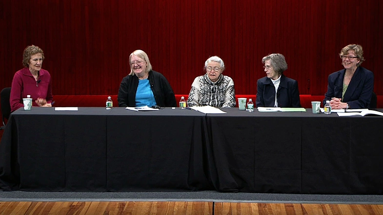 Five speakers on a panel sit at a long black table on stage.