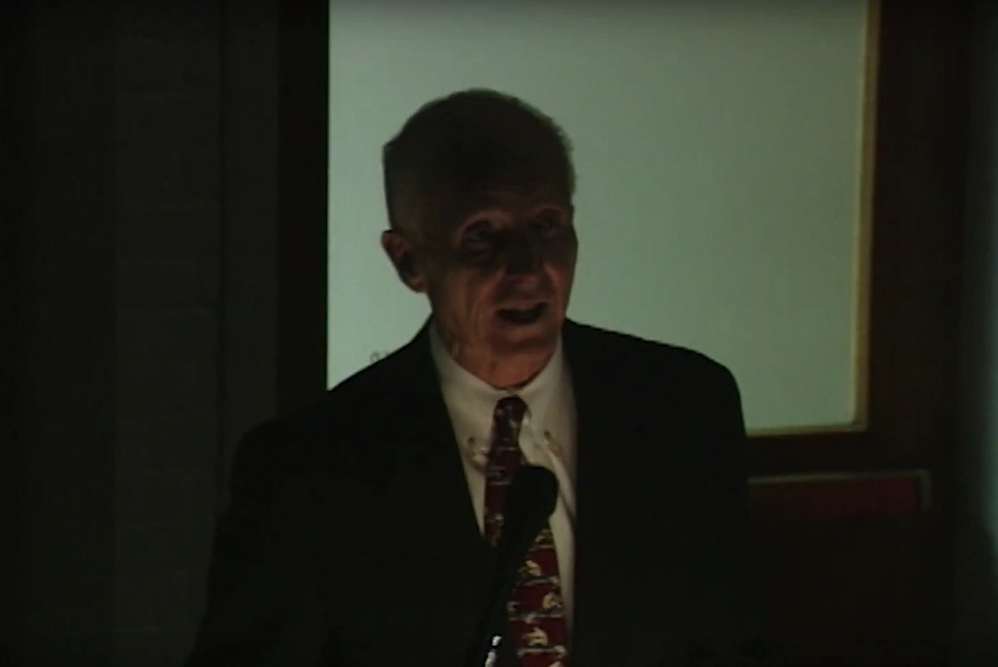 Joe G. Gavin, Jr. speaks into a microphone at podium in a dark room, blank white projector screen behind him.