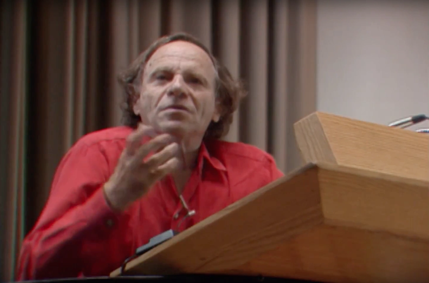 Philip Morrison speaking at podium on stage, gesturing with hand for emphasis, cream colored curtain in background