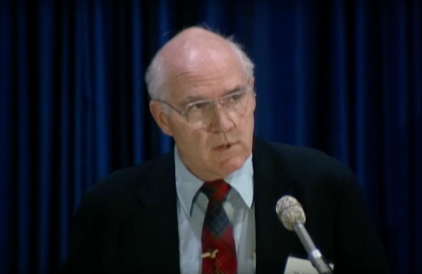William Wallenmeyer speaking at a podium on stage, in front of dark blue curtains