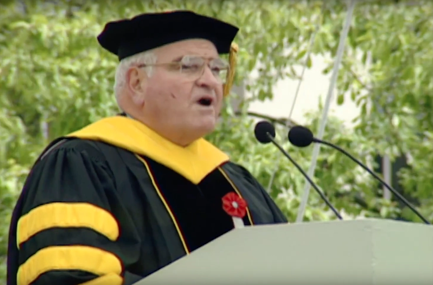 Kaim Aga Khan IV wearing commencement attire, speaking at podium on outdoor stage, trees in background