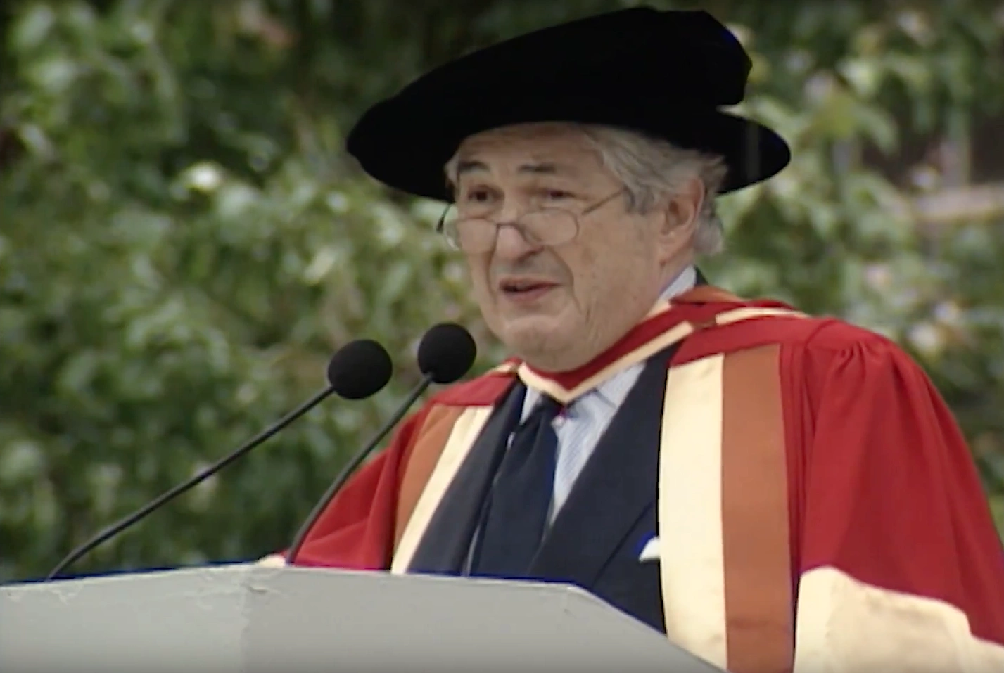 James D. Wolfensohn, wearing commencement attire, speaking at podium on outdoor stage, trees in background