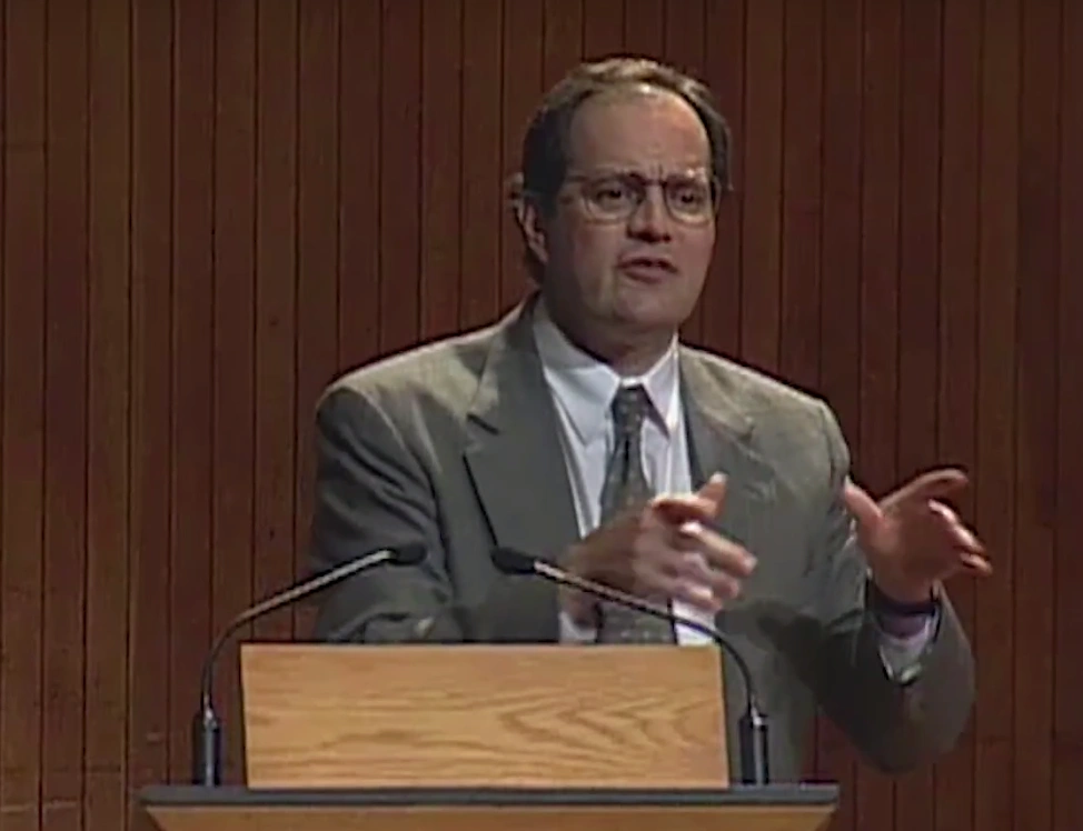 Bob Laughlin speaking at a podium on stage, in front of a red curtain.