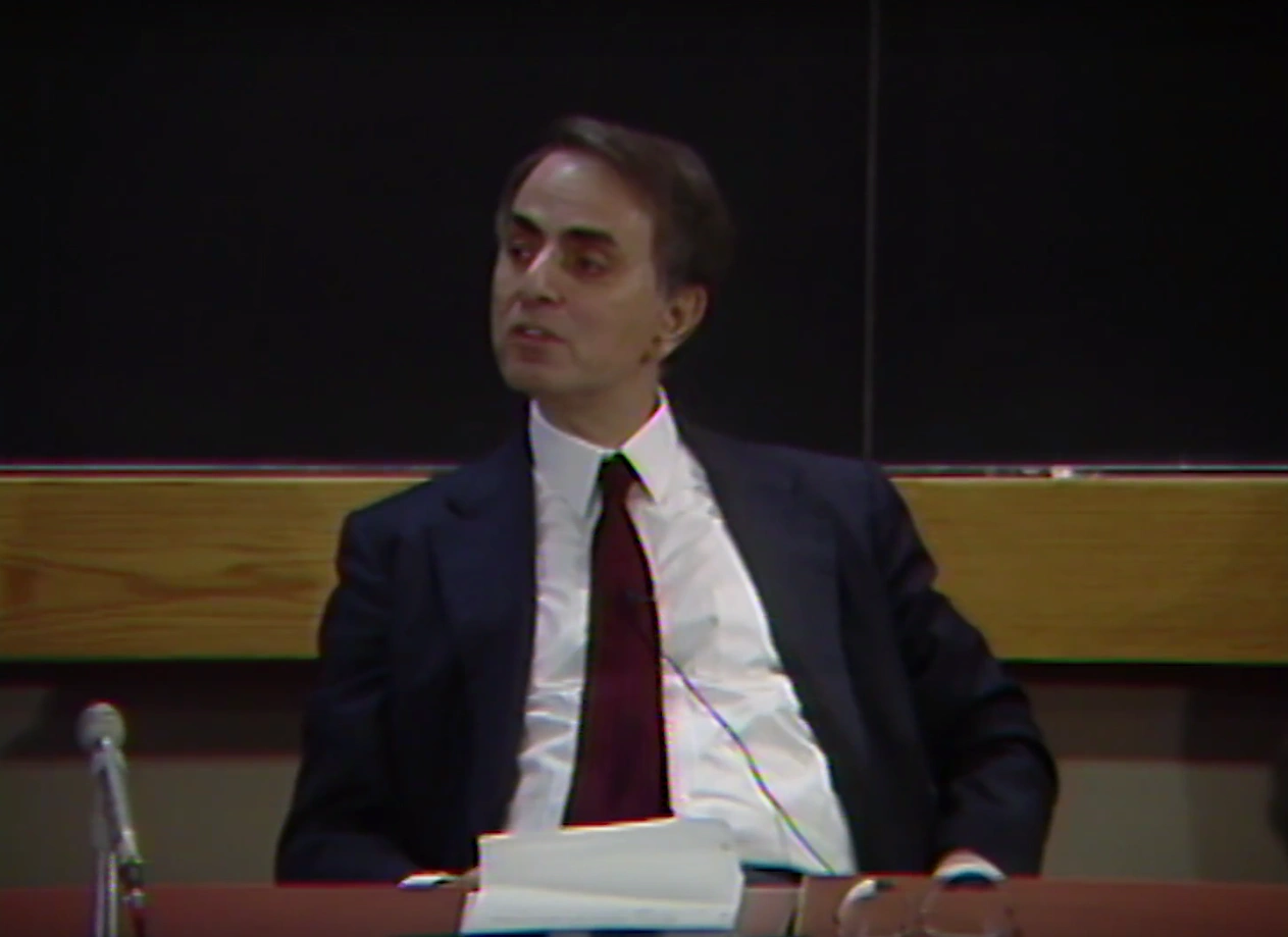 Carl Sagan sits at panelist table in classroom, leaning back in his chair, chalkboard in the background