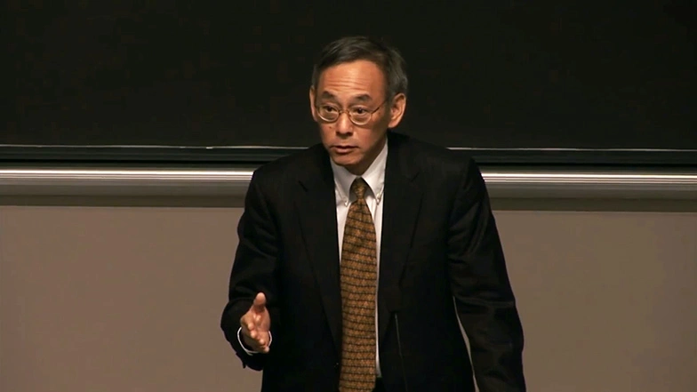 Steven Chu speaks into podium microphones in front of a chalkboard