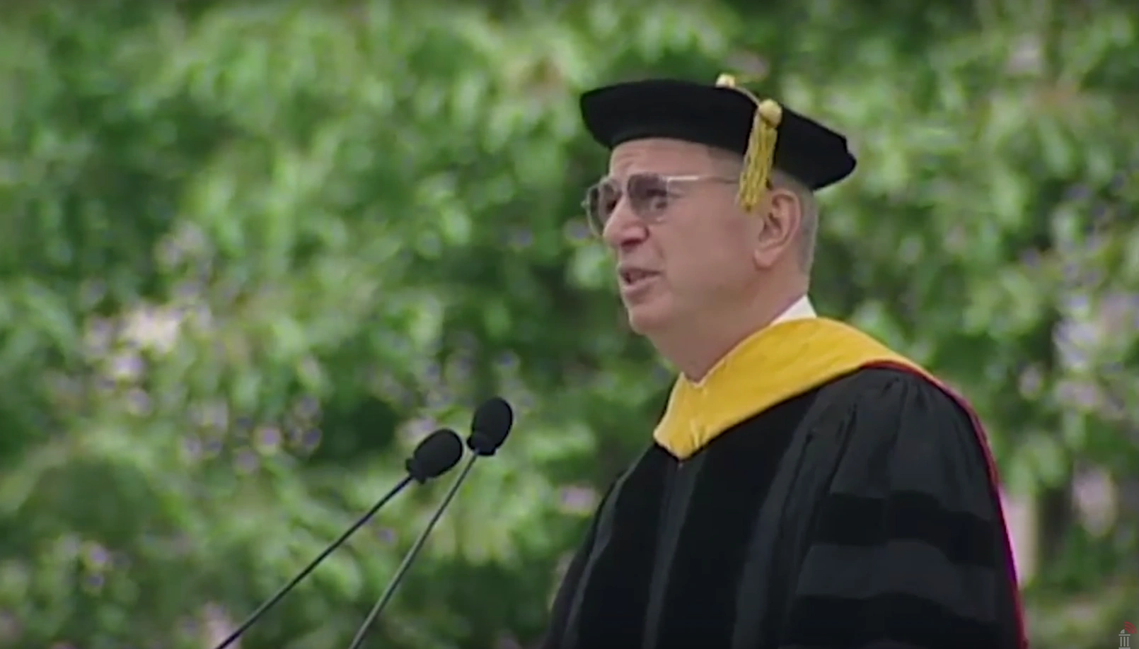 Irwin M. James, wearing commencement attire, speaking at podium on outdoor stage, trees in background