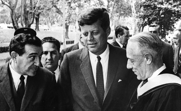 President John F. Kennedy chatting with Jerome Wiesner and Walt Whitman Rostow outdoors, trees in background.