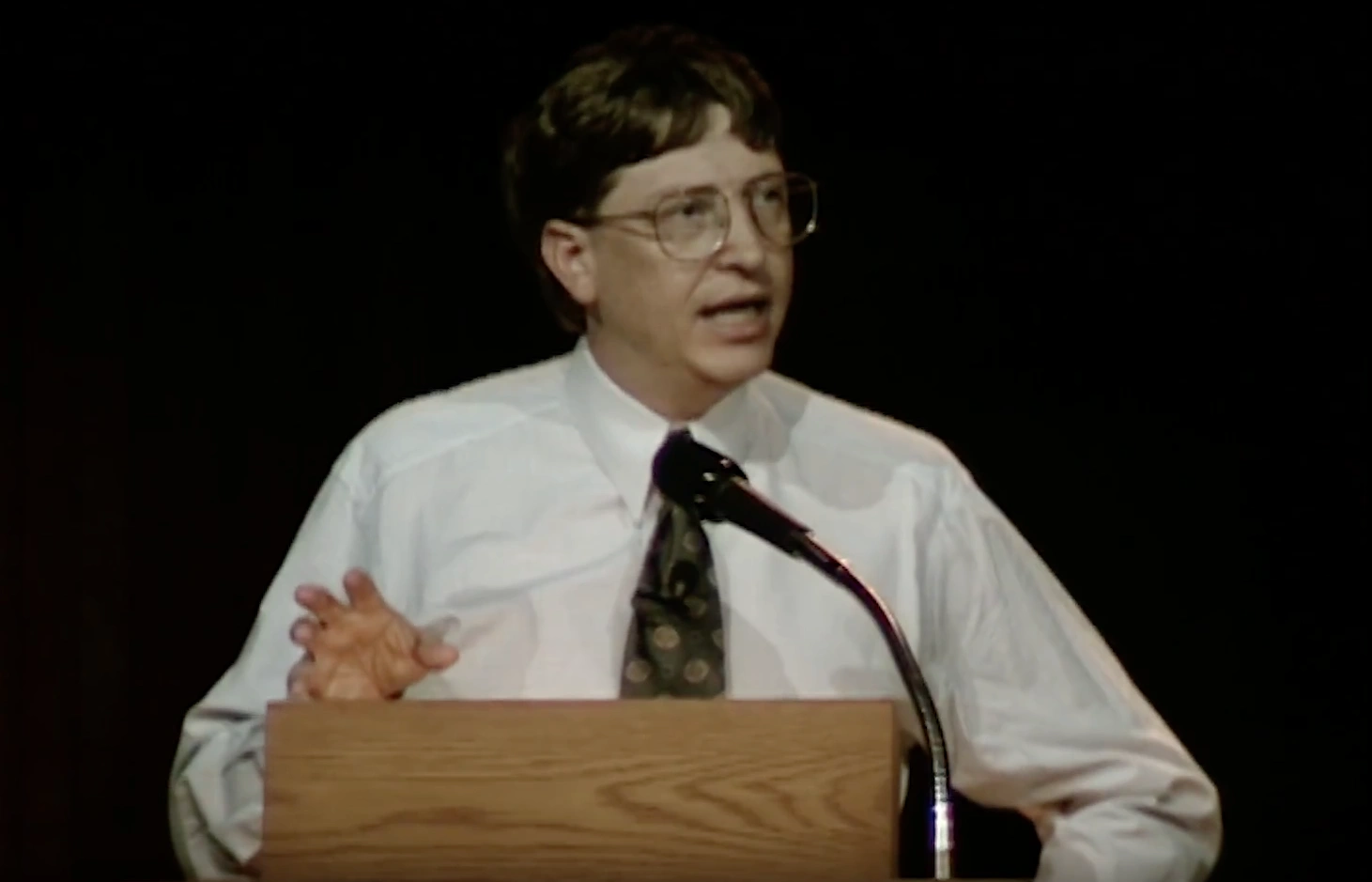 Bill Gates speaking at a podium on stage, in front of a dark background