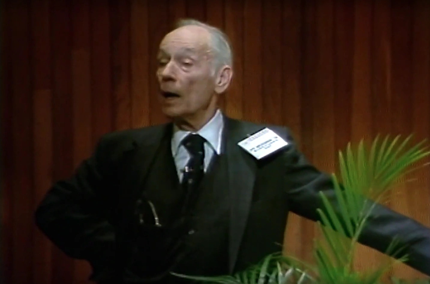  Herman Mesiner speaks at a podium onstage, red curtain in background and decorative palm frond beside him