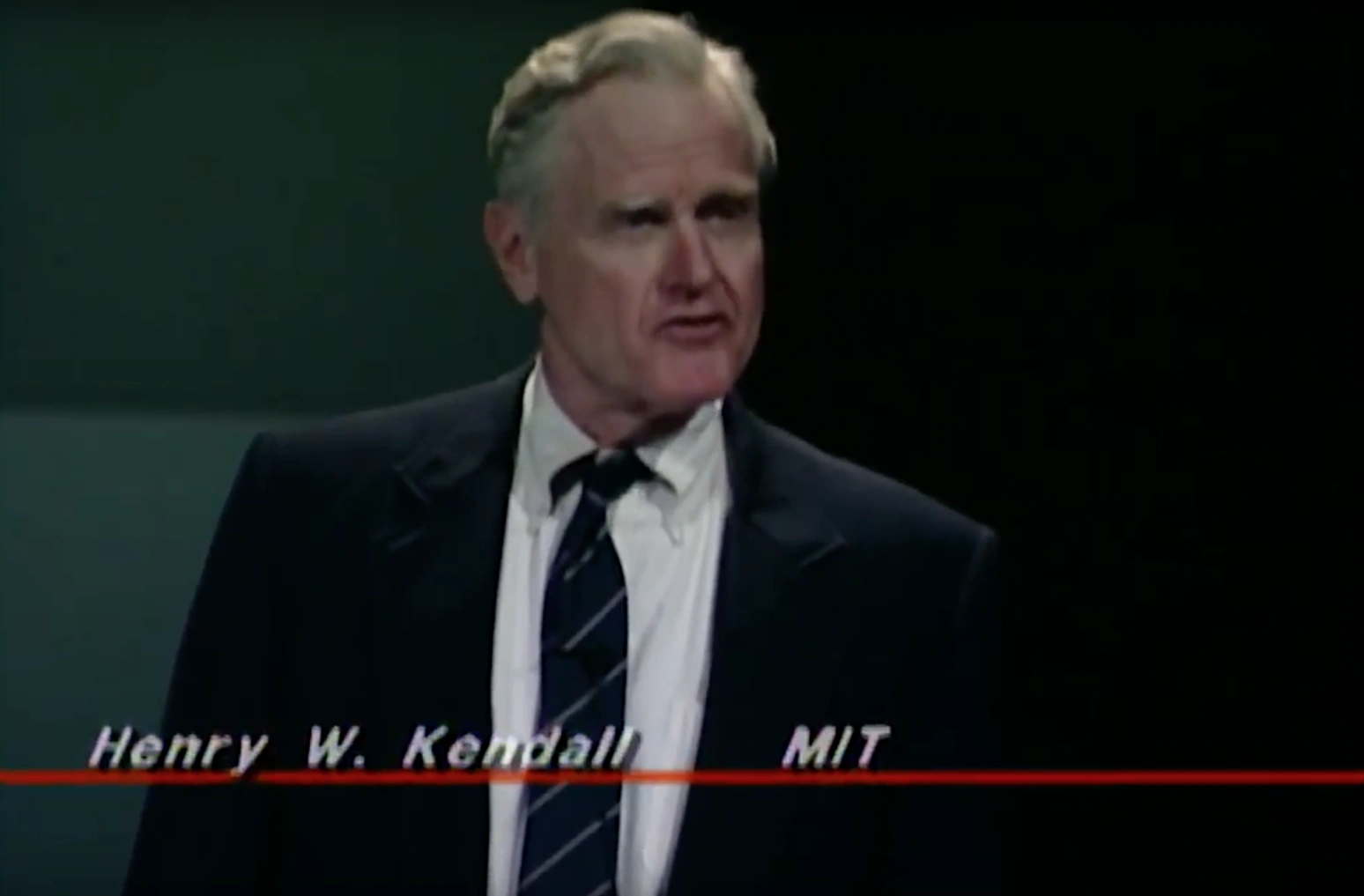 Henry W. Kendall standing on stage and speaking, dimly lit green walls in background