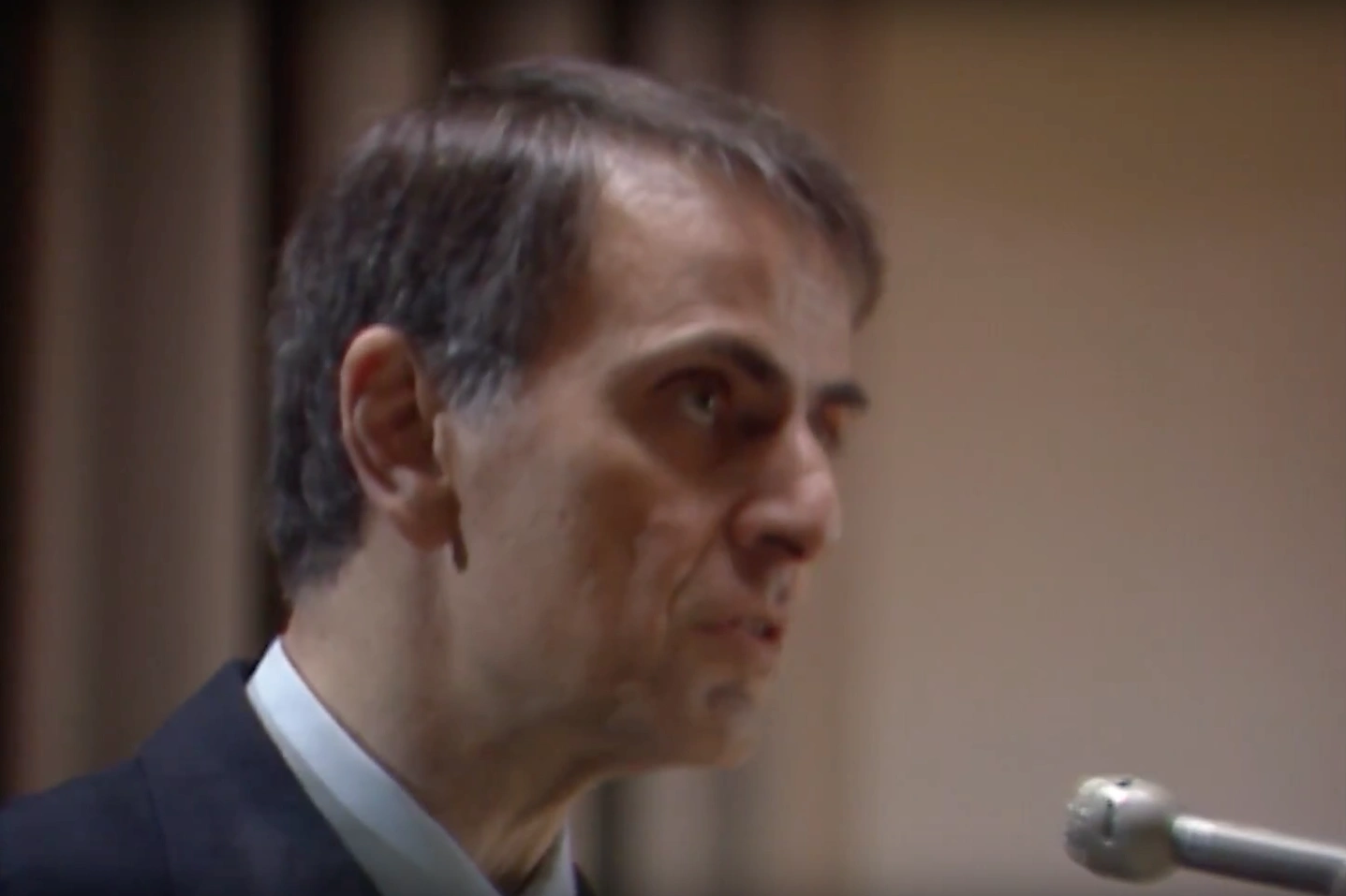 Carl Sagan staring forward as he speaks at a podium, cream curtains and walls in background.