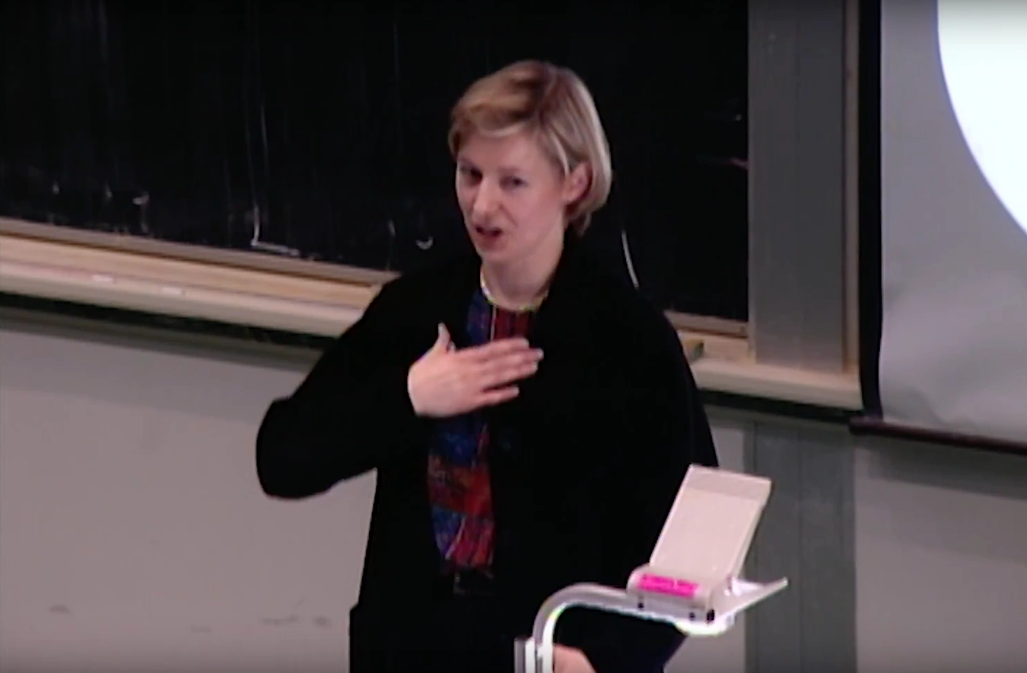 Rosalind Picard lectures in classroom, placing a hand on her heart, chalkboard and projector screen in background 