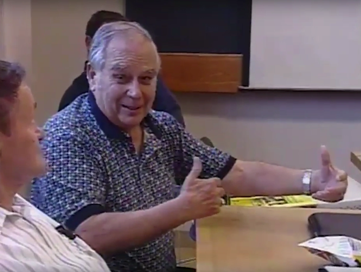 Man sits at table in classroom for Apollo Guidance Computer Project, expressively speaking to person beside him.