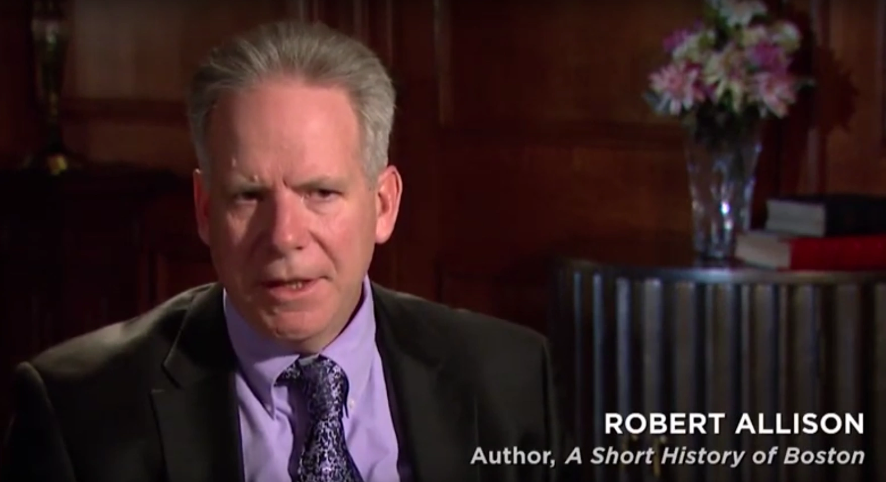 Author Robert Allison seated and mid-sentence in on camera interview, end table with vase of flowers in the background