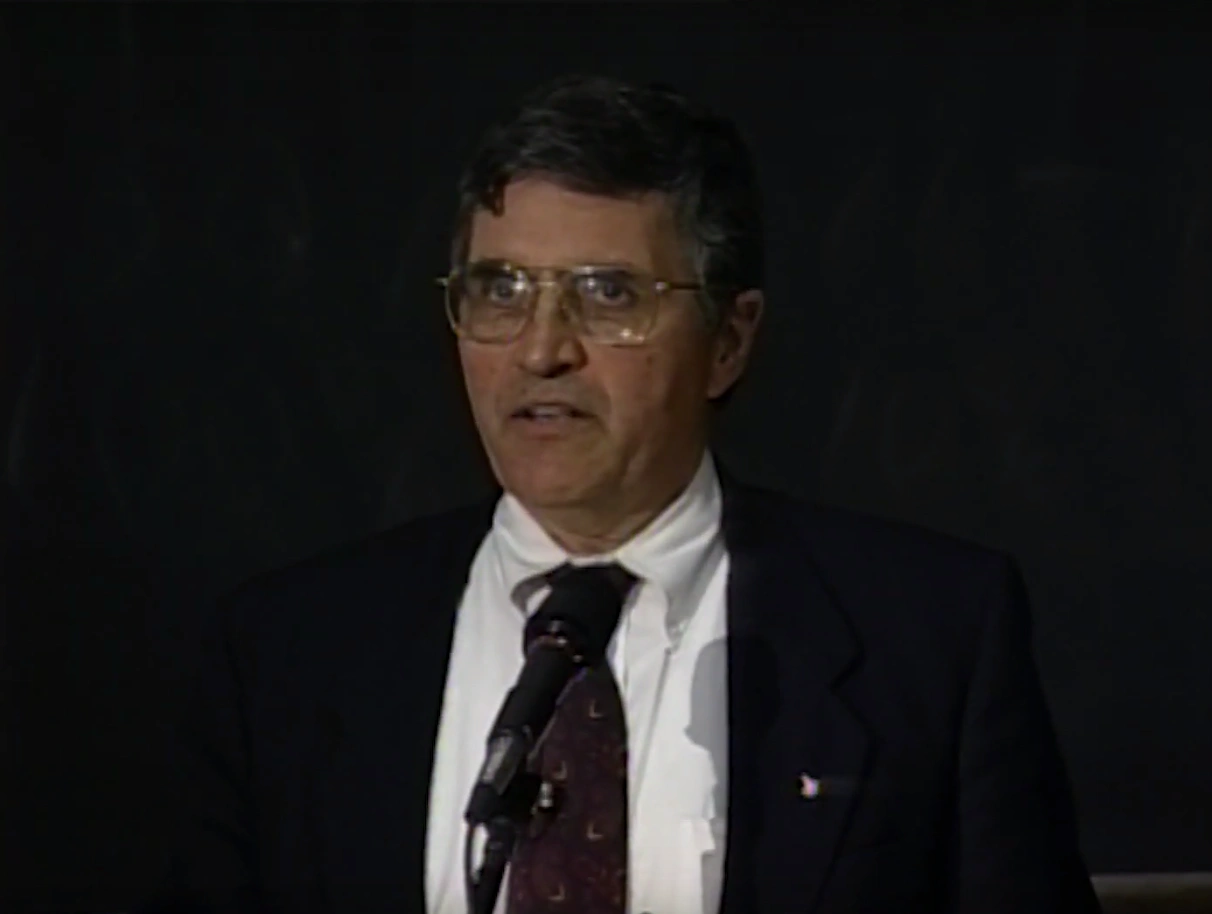 Harrison Schmitt speaking at a podium on stage, in front of a dark background