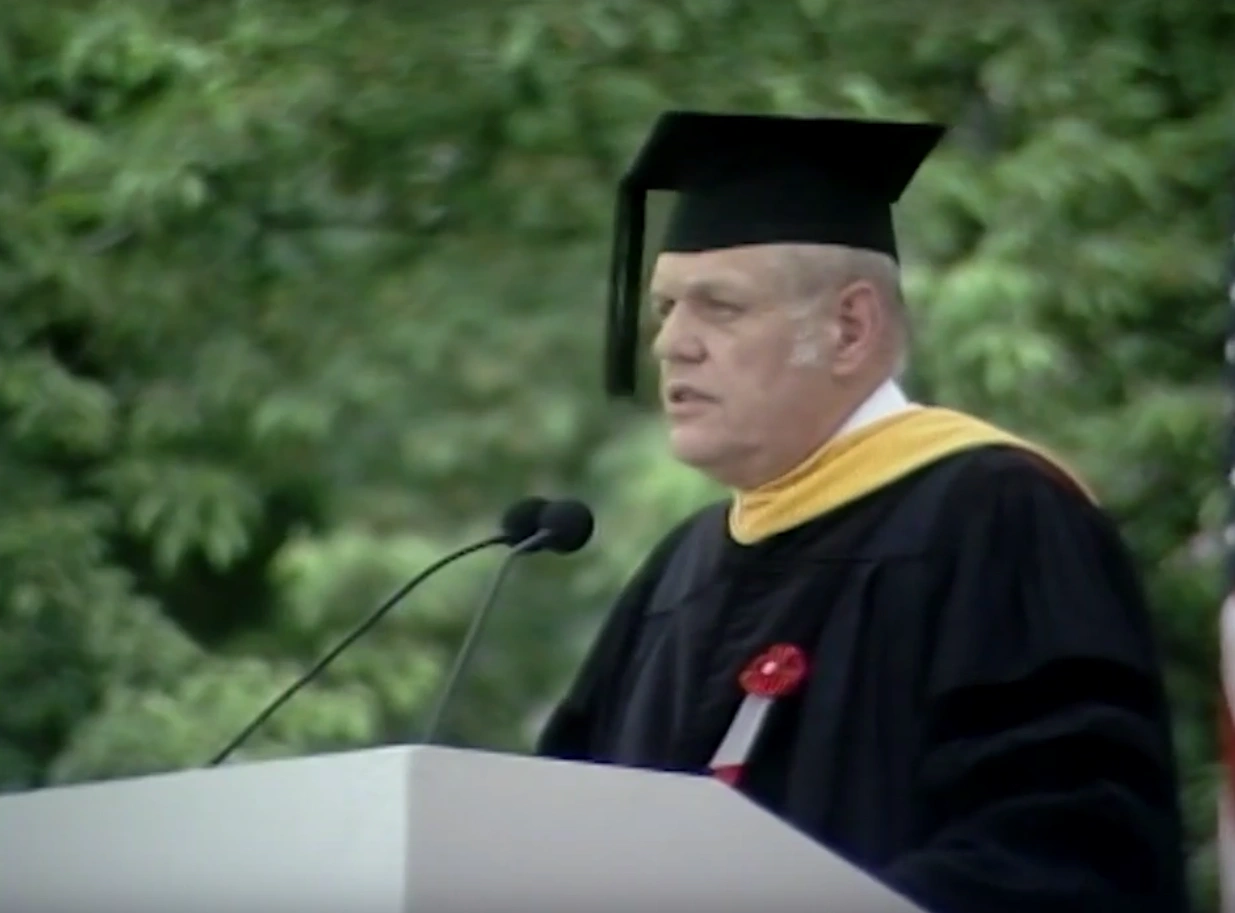 Kenneth Olsen, wearing commencement attire, speaking at podium on outdoor stage, trees in background