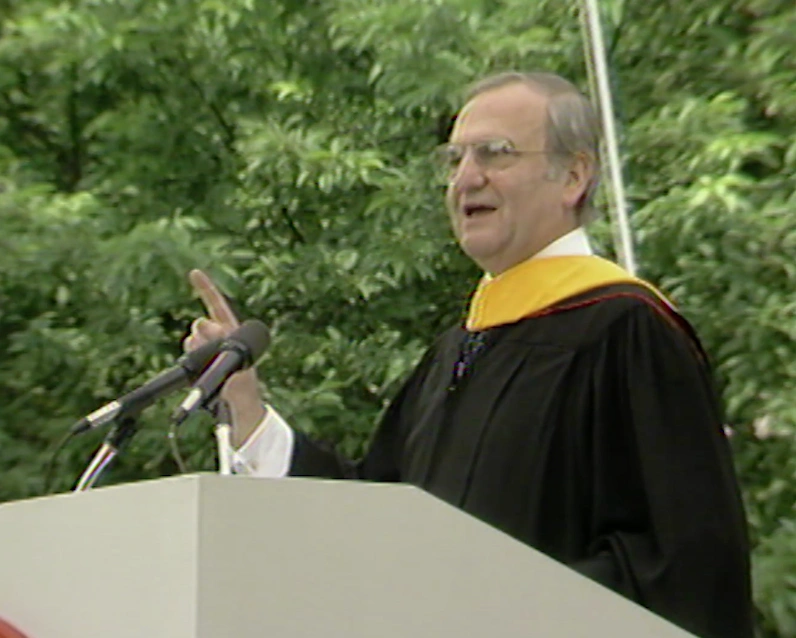 Lee Lacocca, wearing commencement attire, speaking at a podium on an outdoor stage, trees in background
