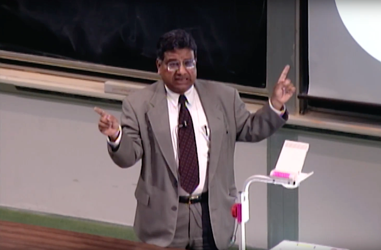 Bijoy Misra lecturing in a classroom, gesturing to projector screen behind them, chalkboard in background.